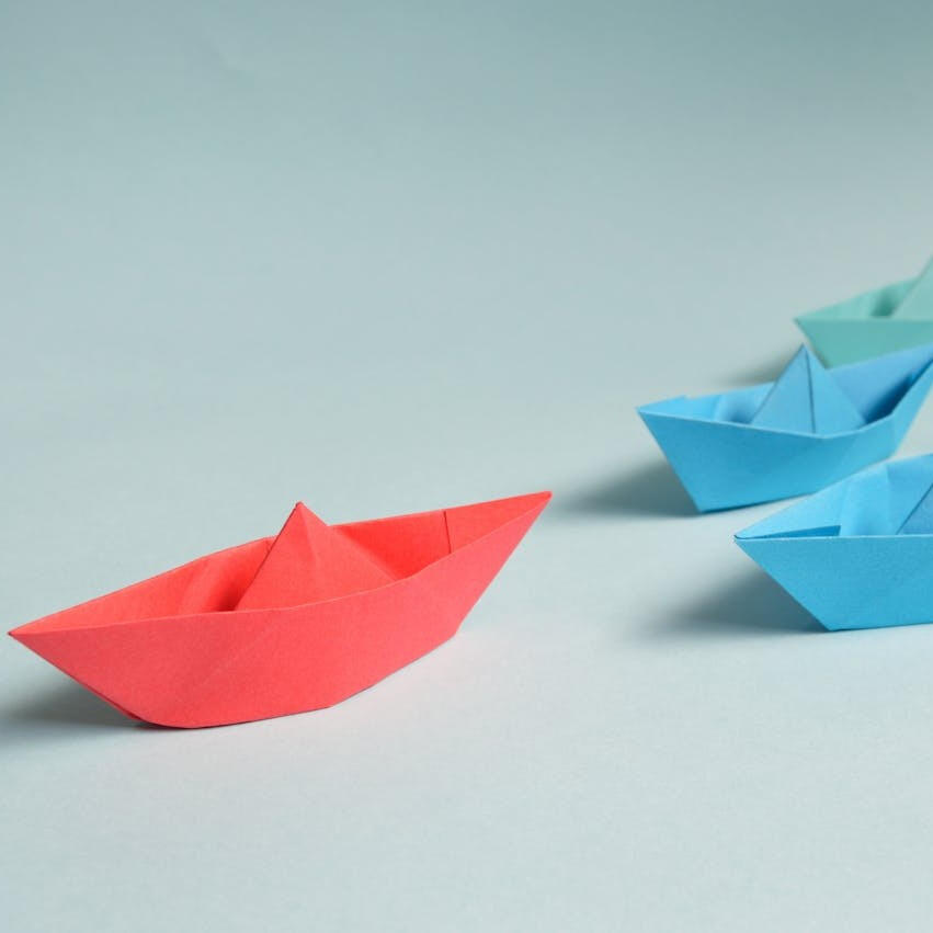 A red paper boat takes the lead in front of three blue paper boats